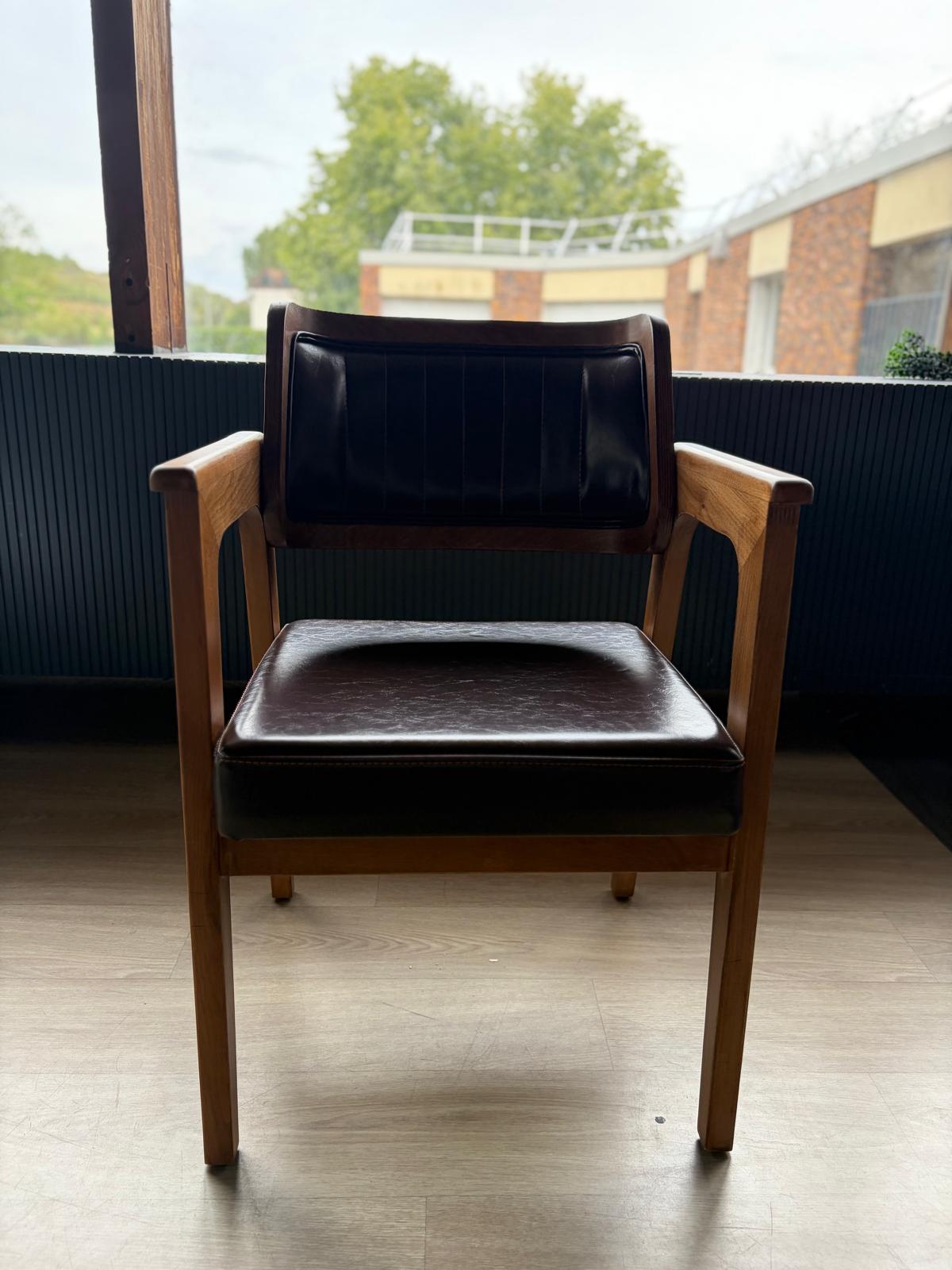 Chaise en bois avec dossier à cadre foncé et assise en simili cuir marron à surpiqûres verticales, vue de face devant une fenêtre
